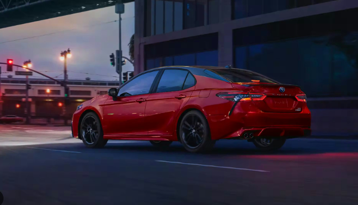 A red Toyota Camry Hybrid is waiting at a red light in an urban area at dusk